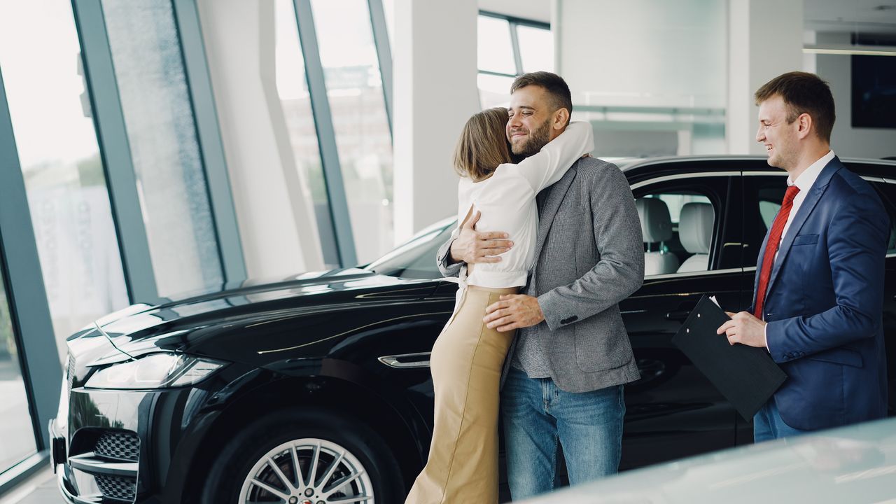 Home Couple hugging with car salesman after purchasing a new car at a dealership.